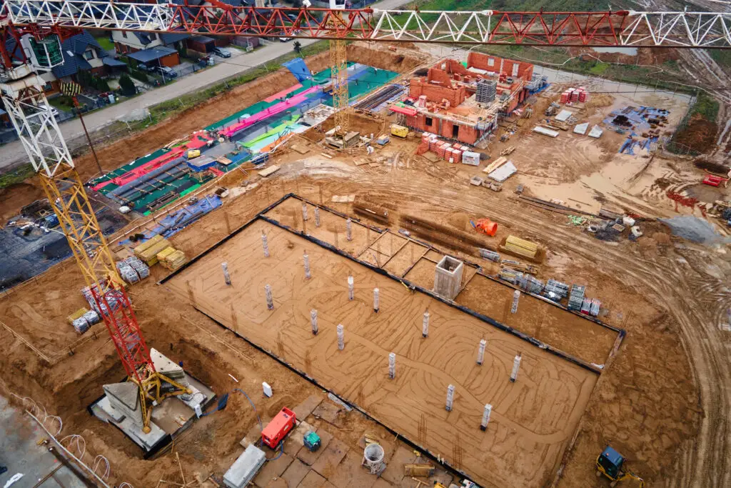 Aerial view of a construction site with a tall crane, a brick building under construction, and scattered materials on muddy ground.