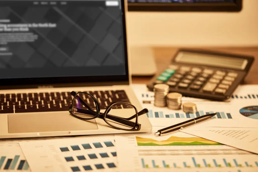Close-up of a laptop, reading glasses, a calculator, and stacks of coins on a desk with financial charts in the background.